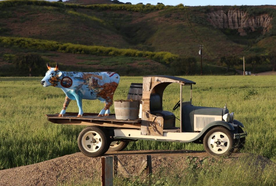 A sculpture of a decorative blue cow on the back of a vintage truck that is sitting on a mound in the middle of a field