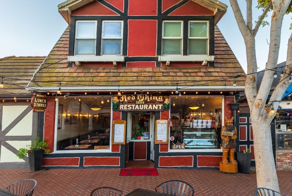 Front facade of a small red restaurant with a brick sidewalk, tall tree and tables and chairs out front