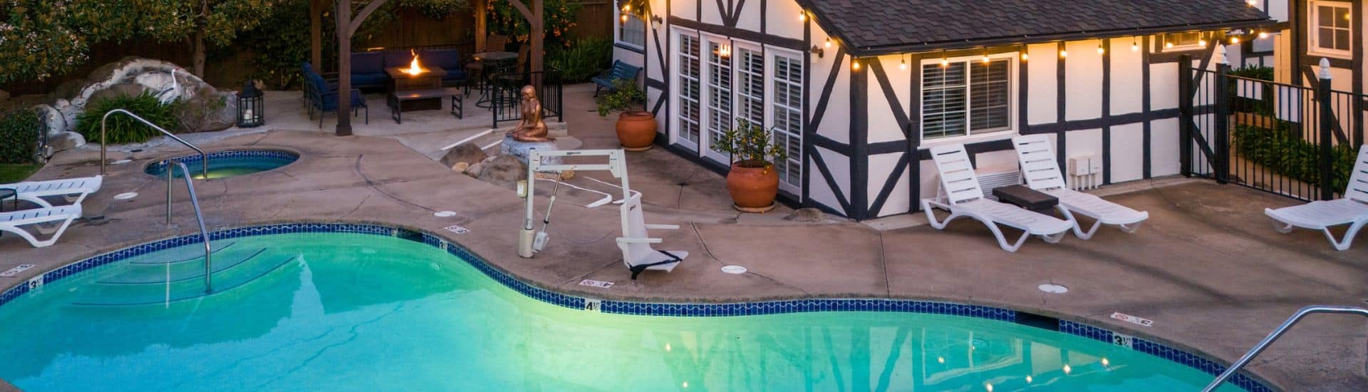 A curved outdoor pool with white lounging chairs, outdoor seating with a firetable, and a white building with brown trim