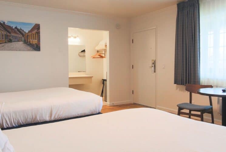 Bedroom with two beds with white sheets and pillows. table with two chairs in front of large window with curtains