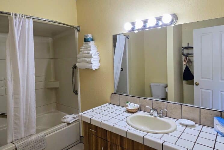 Bathroom with cream walls, tiled vanity with sink, large mirror with bulb lights, shower with white curtain