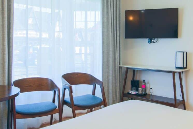 Bedroom with bed, table, two chairs, TV, buffet table and window with sheer and tan curtains