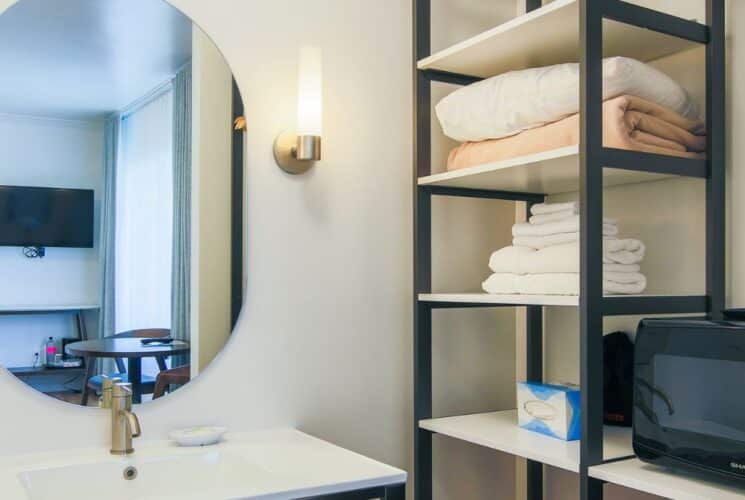 Bathroom with sink. oval mirror, sconces, shelving unit with folded towels and microwave