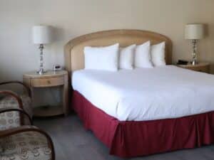 Bedroom with queen bed, two plush sitting chairs, side tables with lamps and bright window with blinds