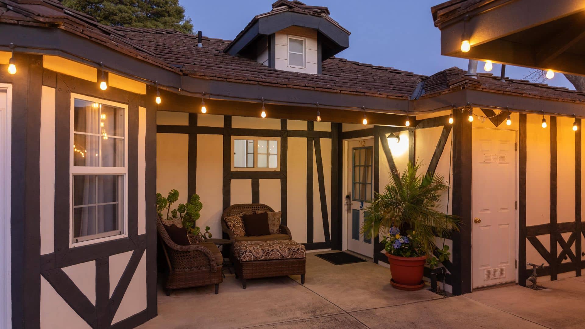 A white building with brown trim, outdoor seating by a front door and patio lights lit up at night